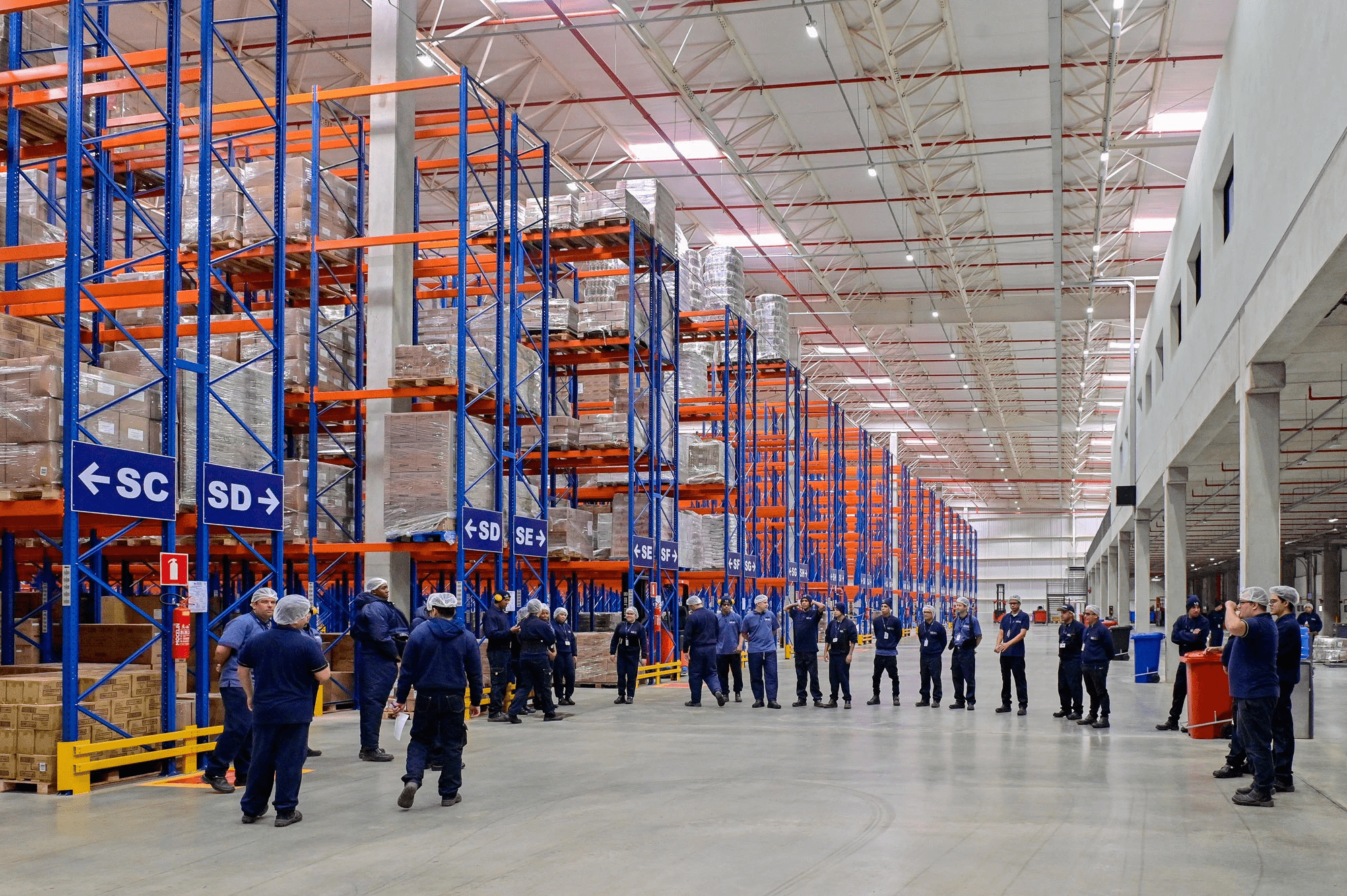 A group of warehouse workers in signature blue shirts gathered inside a massive warehouse for a roll call