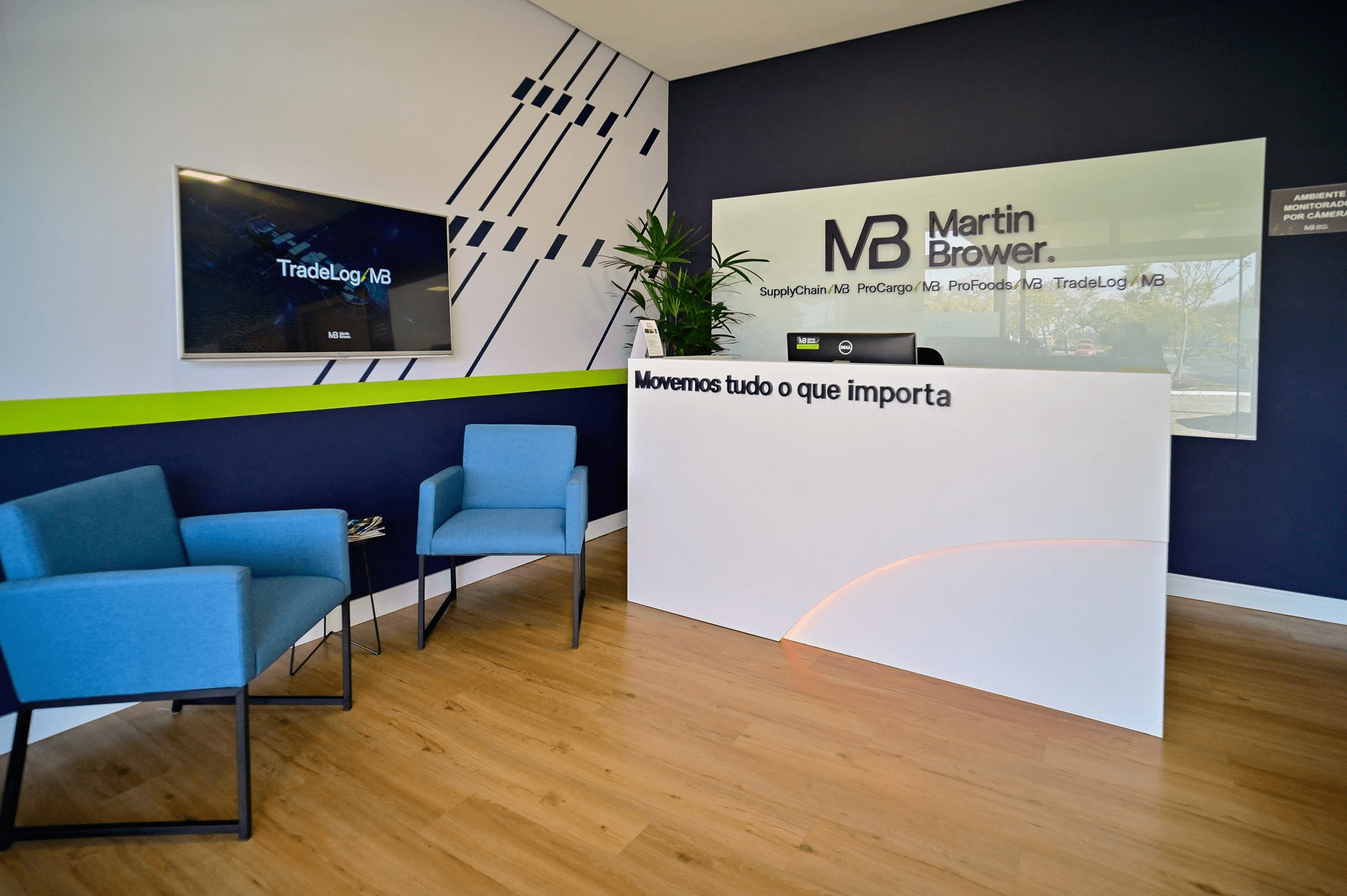 Reception area at the entrance to the production facility. White and blue walls with a green stripe in the center, a TV on the side displaying information about MB Brazil, and a reception desk in the center by the wall