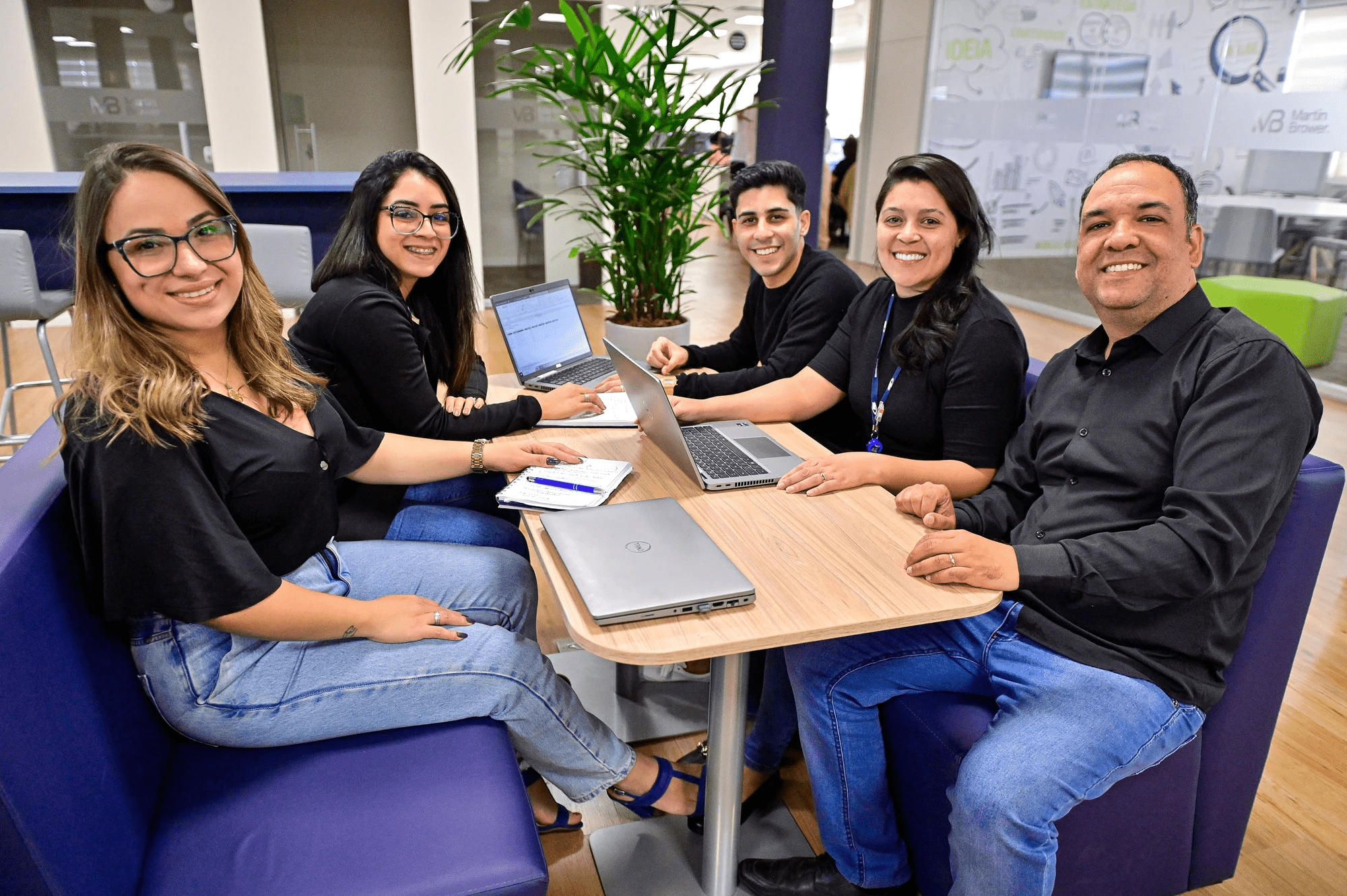 A group of smiling office workers in black shirts and white jeans sitting at a table and working on laptops