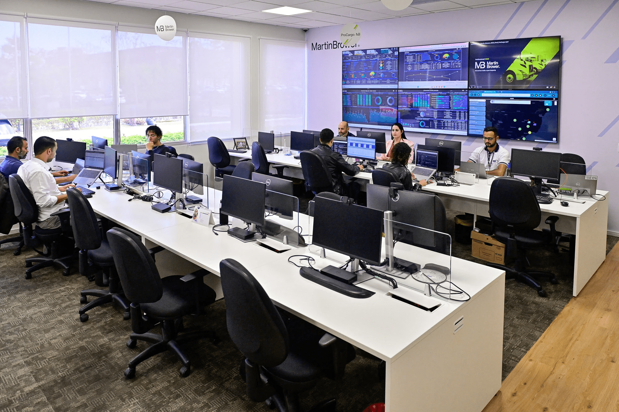 A group of office workers sitting at desktop computers in the monitoring center; on the wall, there are 6 monitors displaying various technical information