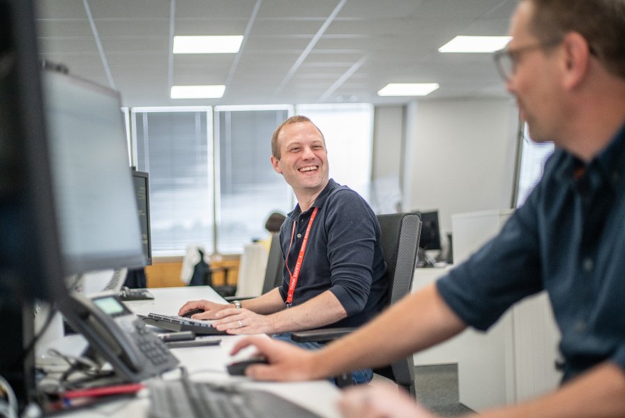 Two laughing men talking to each other seated at desks in an office