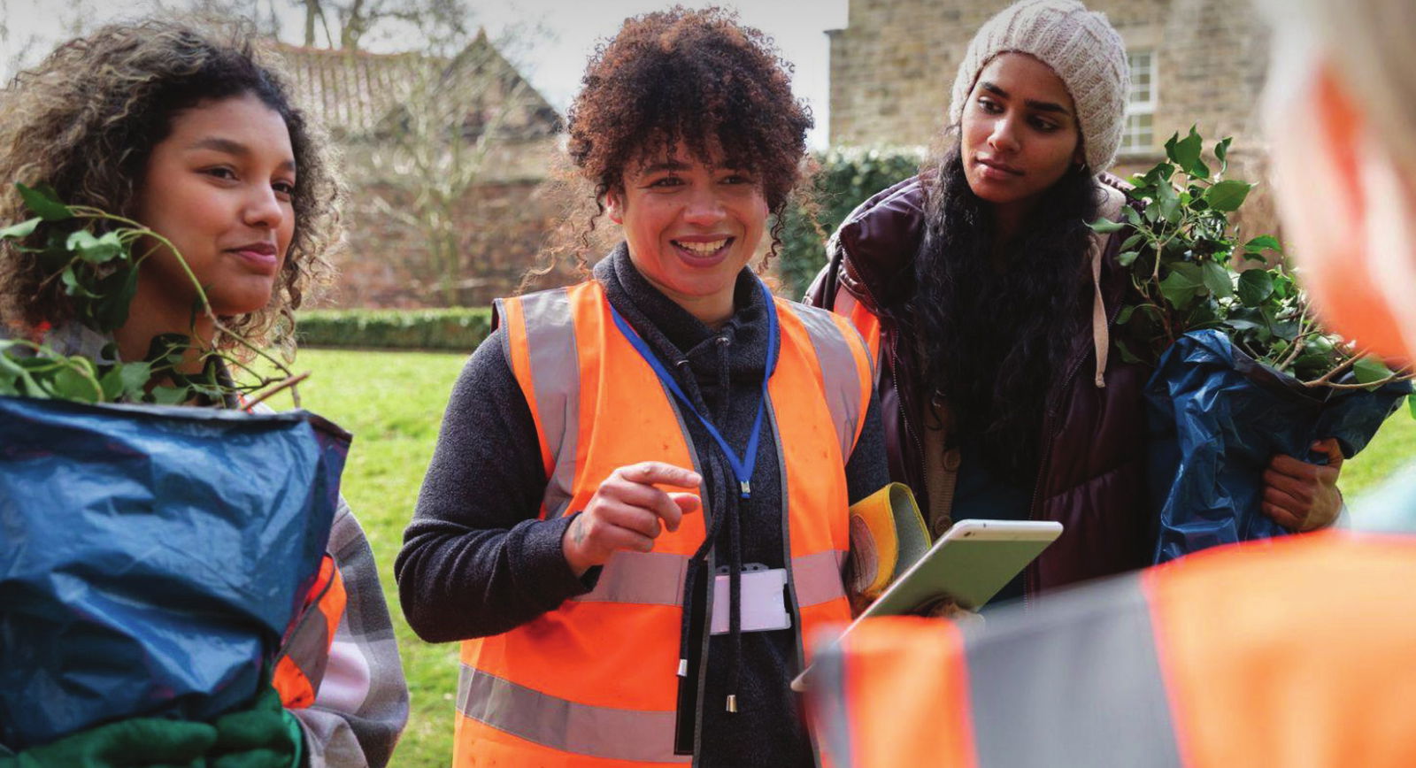 Three women, one wearing an orange vest, carrying plant scraps outdoors with buildings in the background