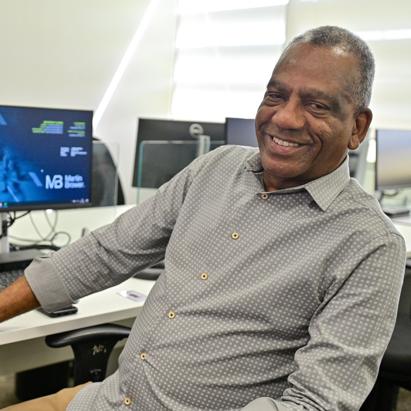 A man smiles while sitting on a chair, turning away from the computer desk