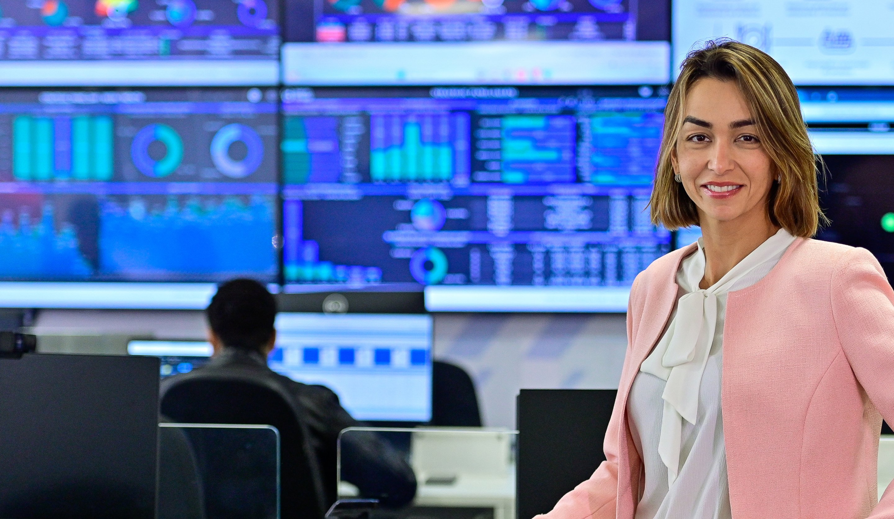 Woman in pink clothes smiling, standing in front of the command center with a lot of computer screens