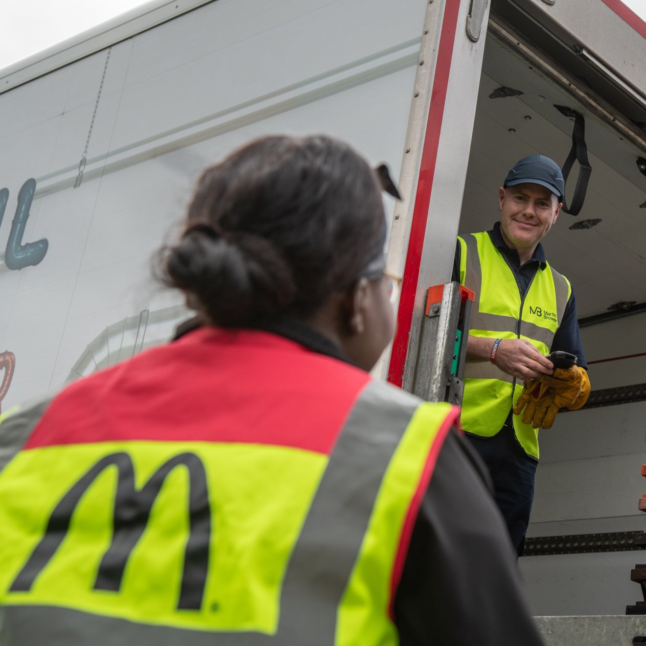 Man wearing a yellow vest standing in the back of a semi truck talking to a woman who is standing below him
