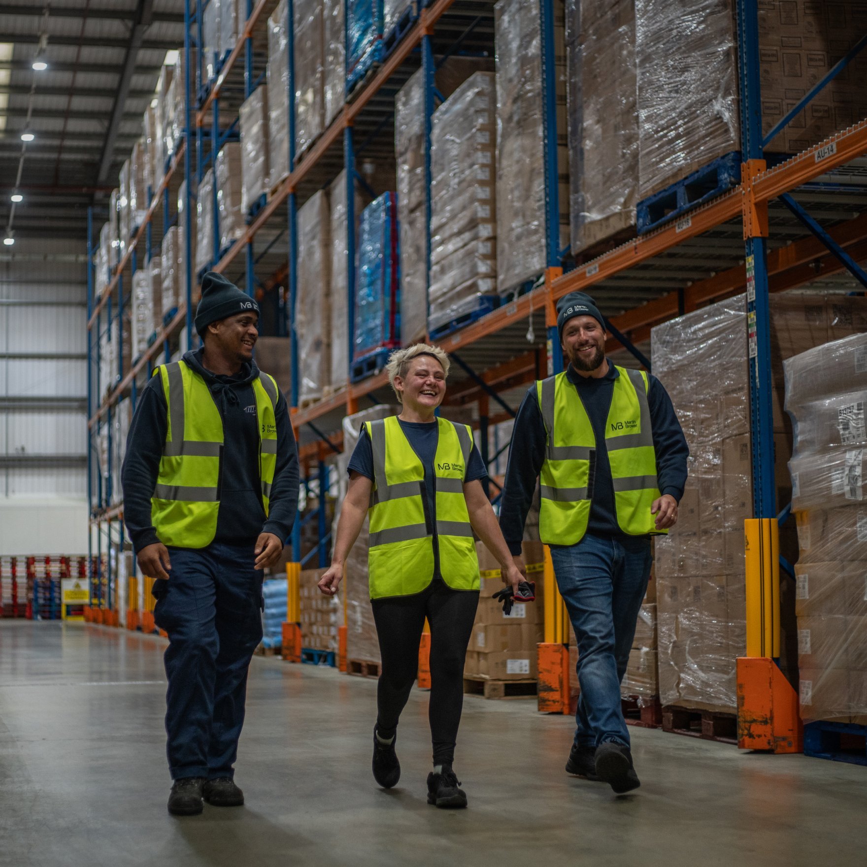 Three people in yellow vests smiling, walking and talking inside a distribution center
