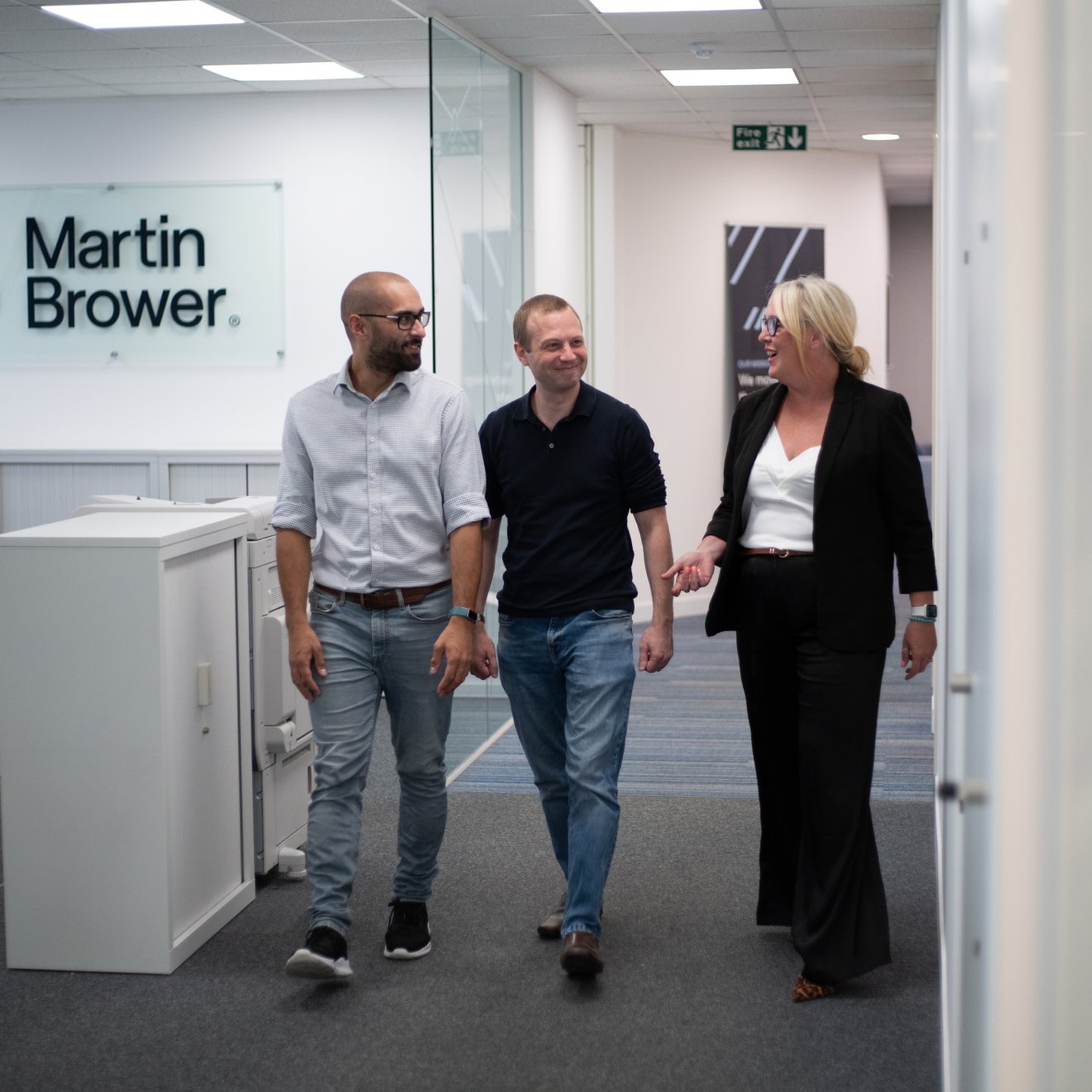 Two men and a woman walking down an office hallway talking and smiling