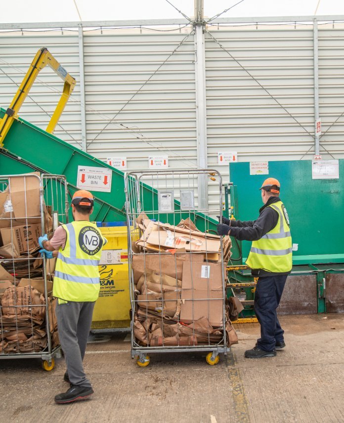 Two men in yellow vests organizing cardboard recycling in front of a green waste bin